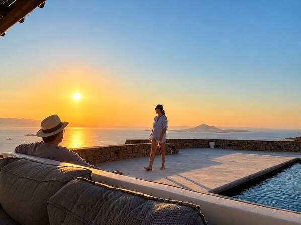 A sunset view from a balcony with a pool, stone walls, and a person standing. - 180-degree panoramic view of the sea and Paros. Stunning sunsets! (Plaka)