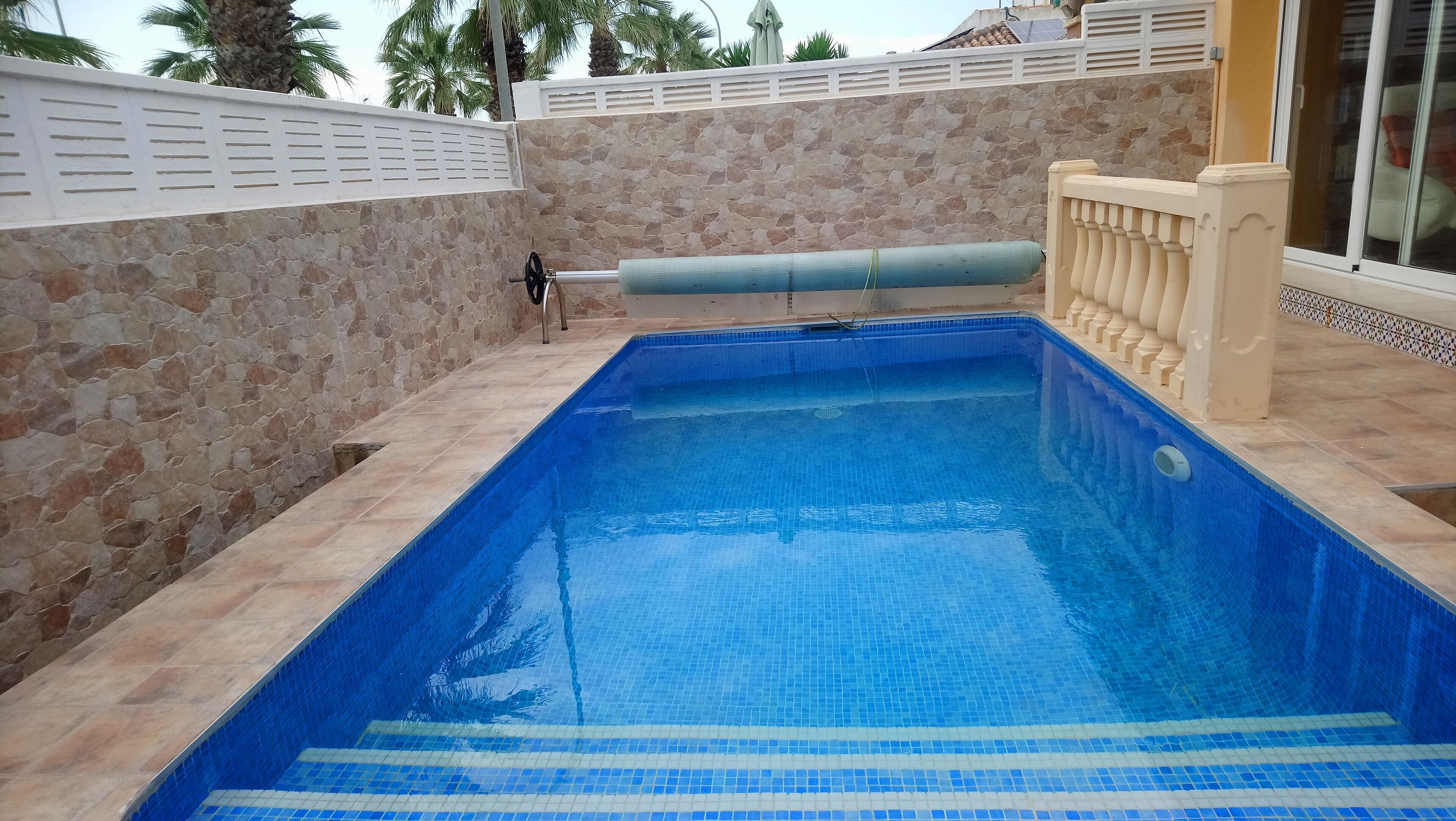 A rectangular swimming pool with blue tiles, surrounded by stone walls and a white fence.