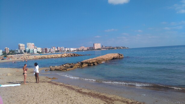 Una platja amb un moll de pedra, aigua blava cristal·lina i un paisatge urbà al fons.