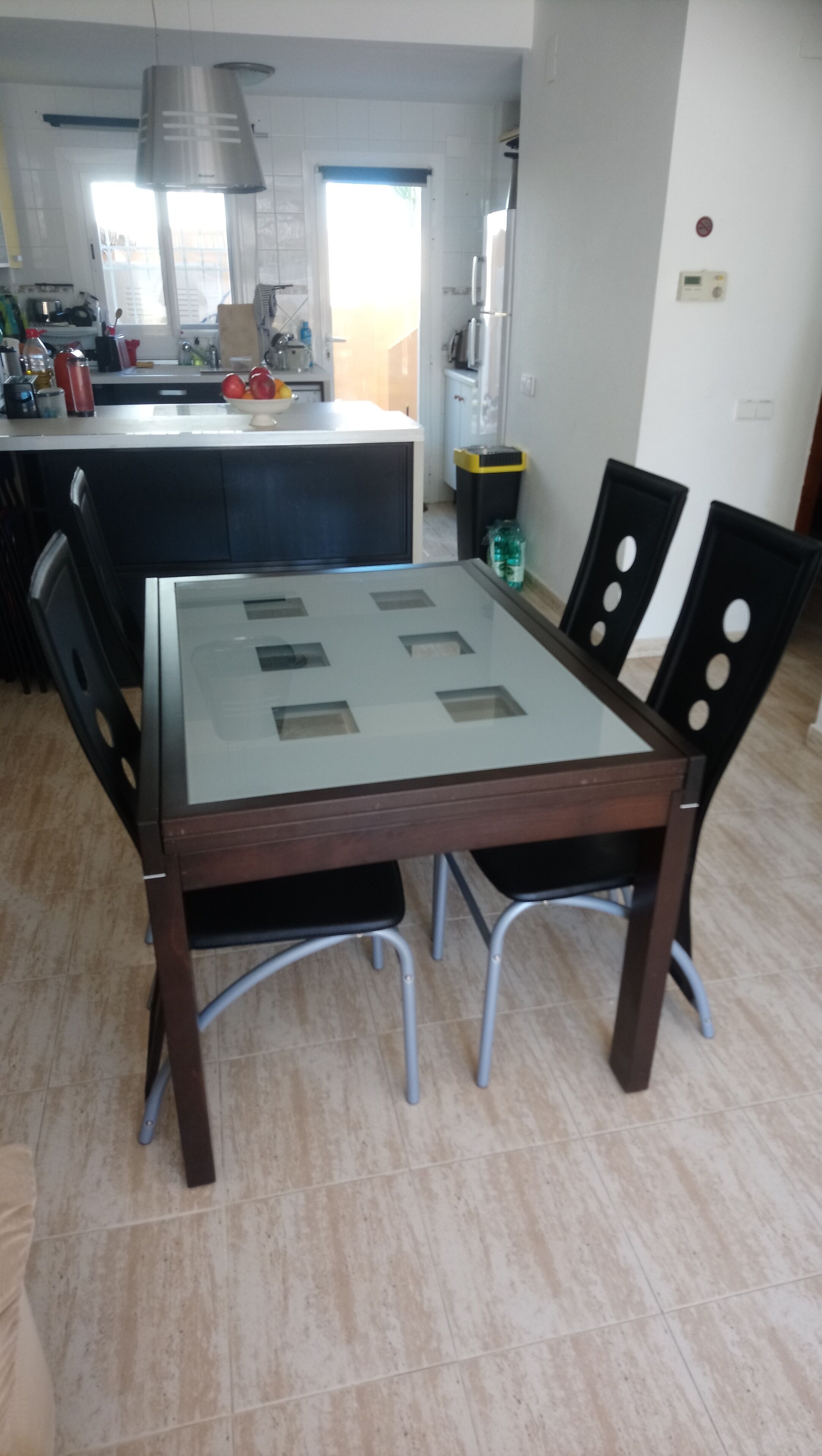 Un comedor con una mesa de vidrio y cuatro sillas, una cocina al fondo y vistas al exterior.