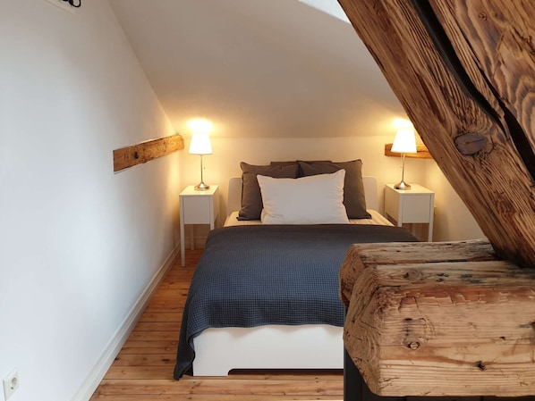 A bedroom with a sloped ceiling, a bed with a blue blanket, two bedside lamps, and a wooden headboard.