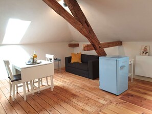 A cozy loft-style room with a wooden floor, a white dining table with chairs, a black sofa with a yellow cushion, and a small blue refrigerator.
