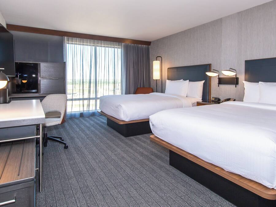 Une chambre d’hôtel avec deux lits, un bureau, une chaise, une télévision et une fenêtre avec des rideaux.