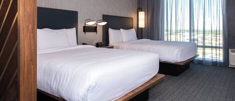 A hotel room with two beds, a wooden headboard, a nightstand with a lamp, and a large window with curtains.
