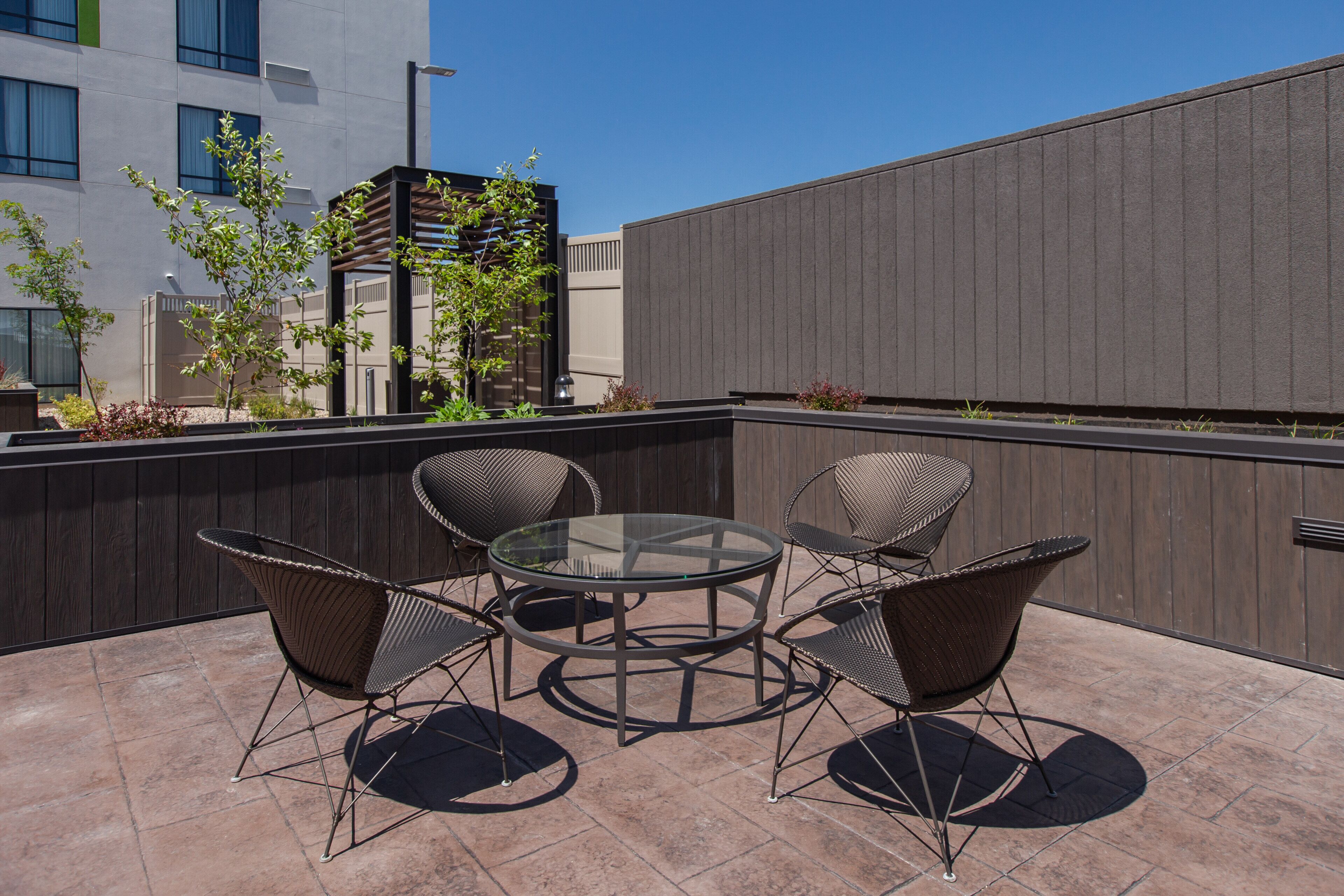 Een terras buiten met een ronde glazen tafel en vier rieten stoelen.