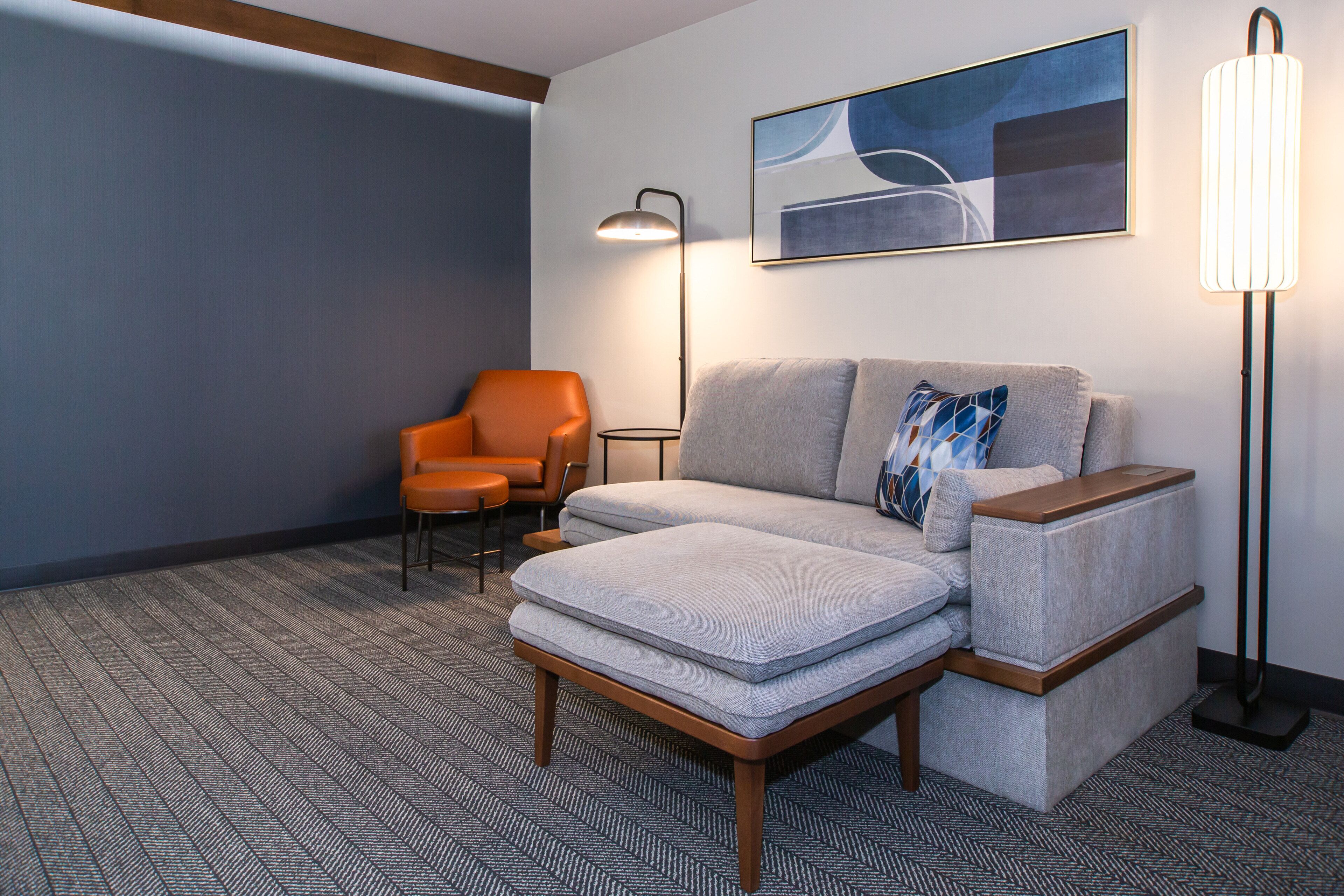 A modern hotel room with a sofa, a chair, a floor lamp, and a painting on the wall.