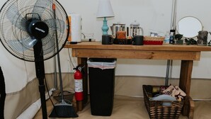 Una habitación con un ventilador de pie, un extintor, un cubo de basura y una mesa con varios objetos, como una lámpara, una cesta y un espejo.