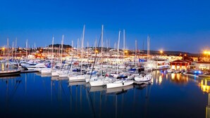 Um porto de iates ao entardecer, com vários barcos atracados, águas calmas e uma cidade distante com luzes.