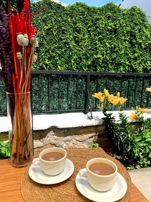 A table with two white cups of tea on a woven placemat, a glass vase with dried plants, and yellow flowers in the background. - Marin Port Otel (Cesme)