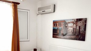 Quarto de hotel com cama, cortinas brancas, um quadro, ar-condicionado e mesa de cabeceira.