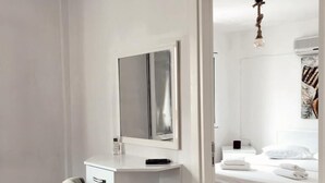 Quarto moderno com chão de madeira, escrivaninha branca com espelho, poltrona cinza e cama bem arrumada com roupa de cama branca.
