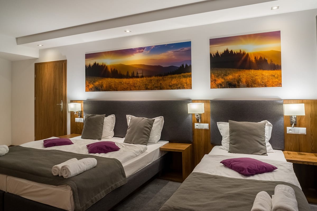 Ein Hotelzimmer mit zwei Betten, jeweils mit Kopfstütze, Nachttischen und Lampen. An der Wand über den Betten befinden sich Gemälde.