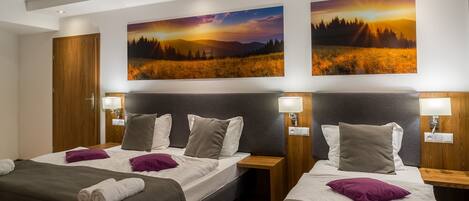 A hotel room with two beds, each with a headboard, bedside tables, and lamps. There are paintings on the wall above the beds.