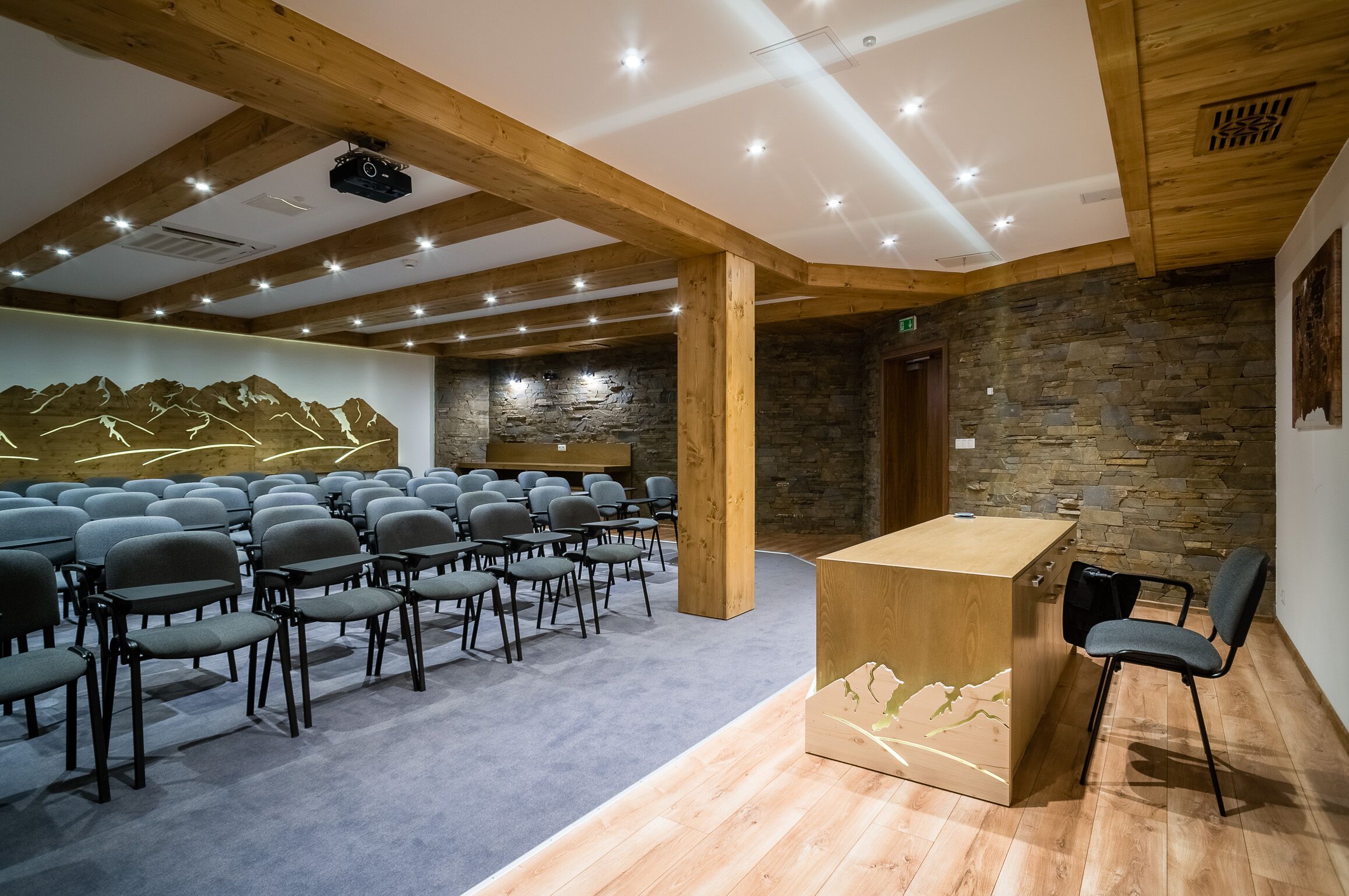Une salle de conférence avec des rangées de chaises, un pupitre en bois et un mur de pierre orné d’une fresque représentant une montagne.
