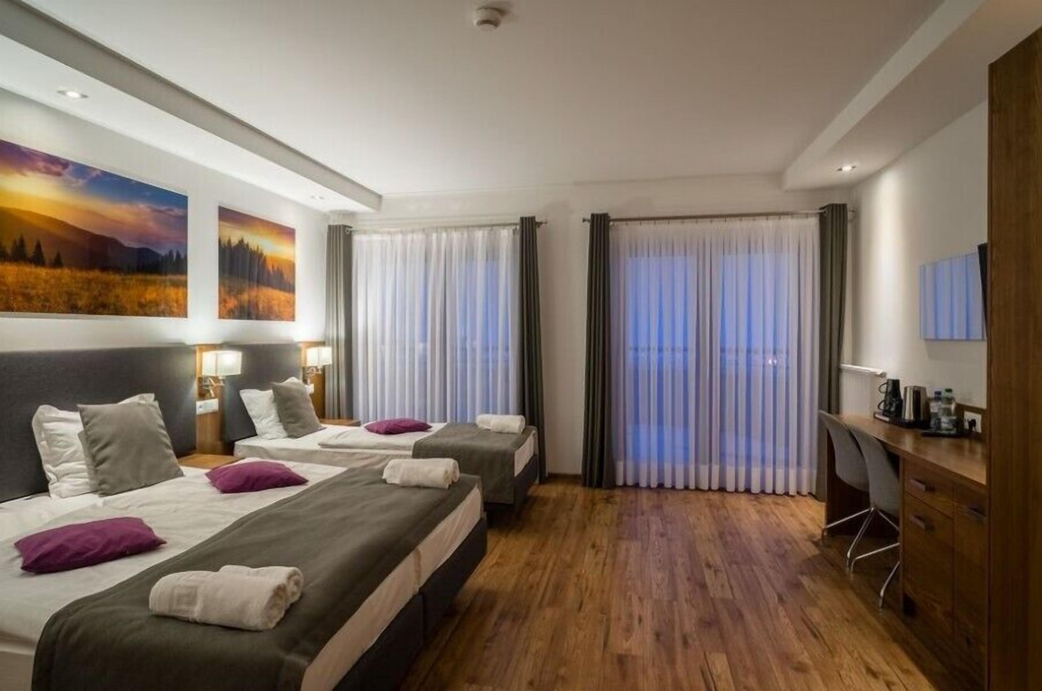 A hotel room with two beds, a desk, and a large window with curtains.