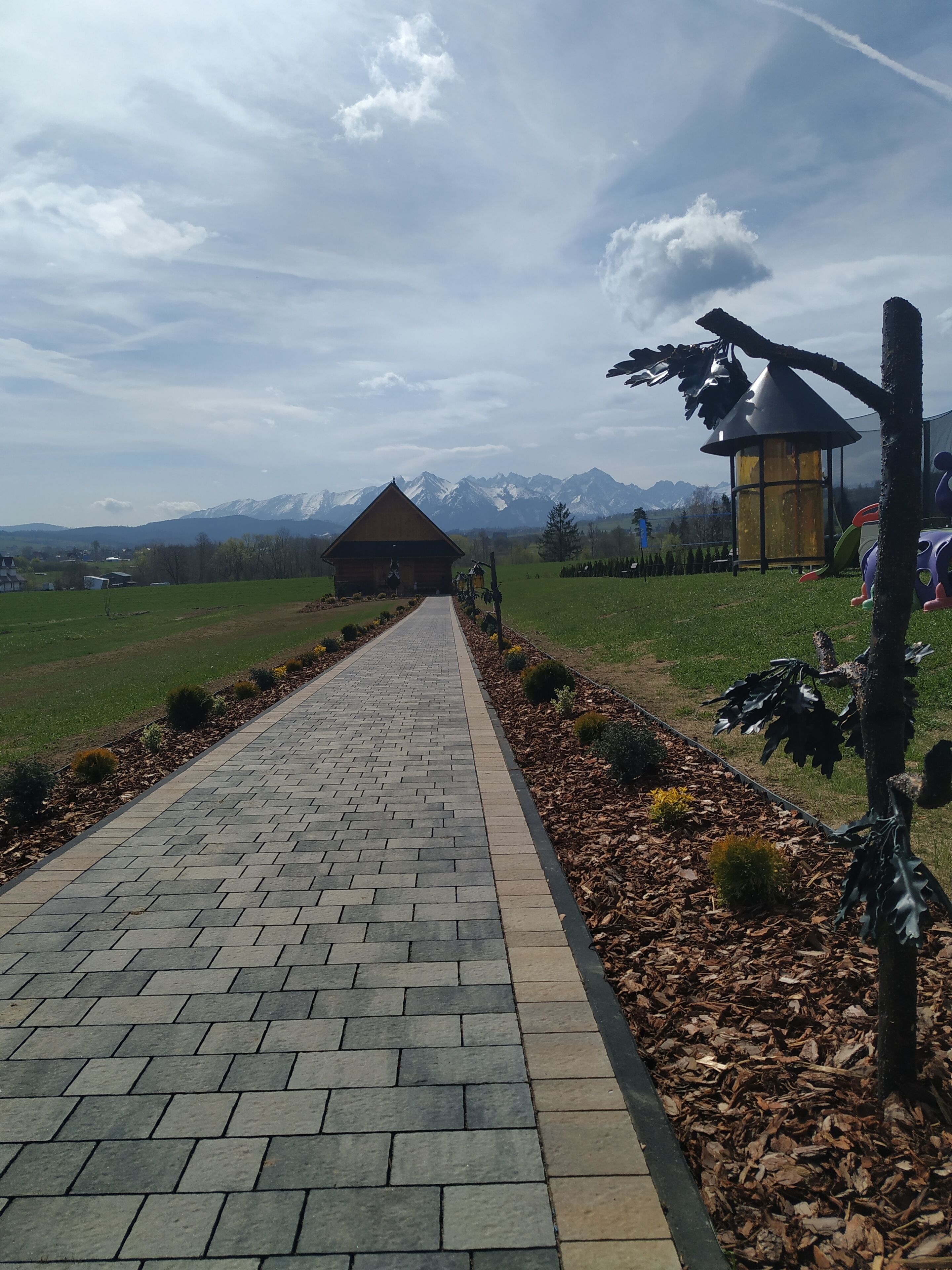 A paved pathway leading to a wooden cabin with a conical roof, surrounded by a landscaped garden with mulch and small shrubs, under a partly cloudy sky with distant mountains.