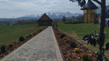 A paved pathway leading to a wooden cabin with a conical roof, surrounded by a landscaped garden with mulch and small shrubs, under a partly cloudy sky with distant mountains.