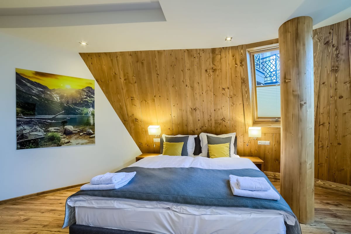 Ein Schlafzimmer mit Holzbalkendecke, einem Bett mit weißer und grauer Bettwäsche, zwei Nachttischlampen und einem Gemälde einer Berglandschaft an der Wand.