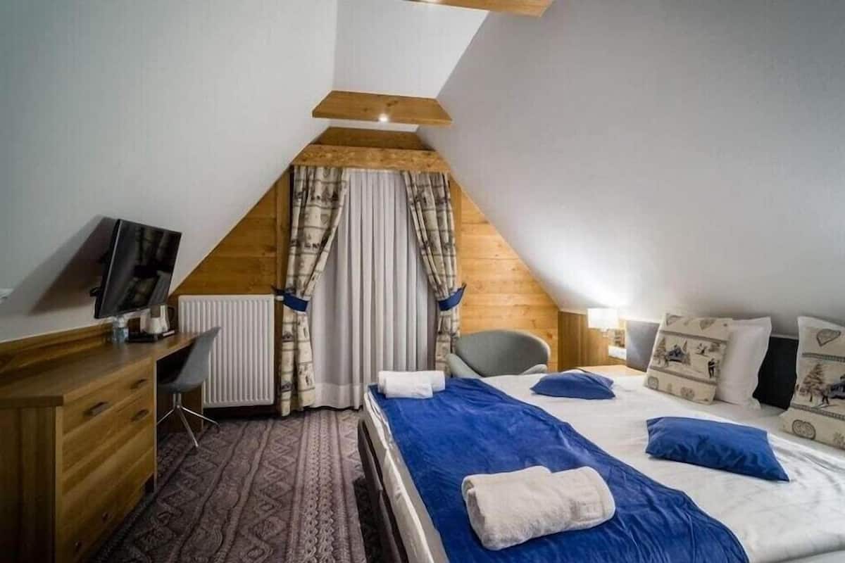 Ein Schlafzimmer mit schrägen Wänden, einem Schreibtisch mit Computer und einem Bett mit blauer Bettwäsche sowie einem Heizkörper.