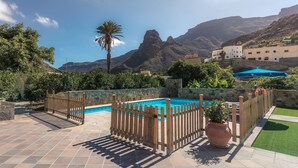 Zona de piscina con valla de madera, una palmera y montañas al fondo.