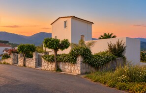 A white building with a small tower, surrounded by stone walls and greenery, with mountains in the background.