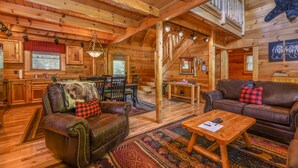 A log cabin interior with a dining area, leather sofas, a wooden coffee table, and a staircase. - FlatBackyard, FirePit, HotTub, Horseshoes, games, 60-game arcade FUN 4 FAMILIES! (Sevierville)