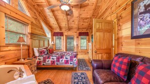 A log cabin interior with a wooden ceiling, a bed with a quilt, a leather sofa, and a bathroom with a sink and mirror. - FlatBackyard, FirePit, HotTub, Horseshoes, games, 60-game arcade FUN 4 FAMILIES! (Sevierville)