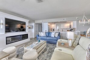 Una sala de estar moderna con chimenea, un sofá azul, un sofá beige, una mesa de centro y una cocina al fondo.