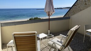 Un balcón con vista a la playa, una silla de madera, una silla blanca, una mesita y una sombrilla.