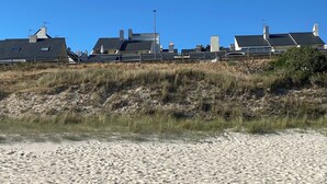Un edificio costero con varios tejados, situado en una playa arenosa con vegetación escasa.