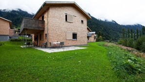Ein zweistöckiges Holzhaus mit Balkon, einer kleinen Terrasse mit Gartenmöbeln und einem üppigen grünen Rasen.