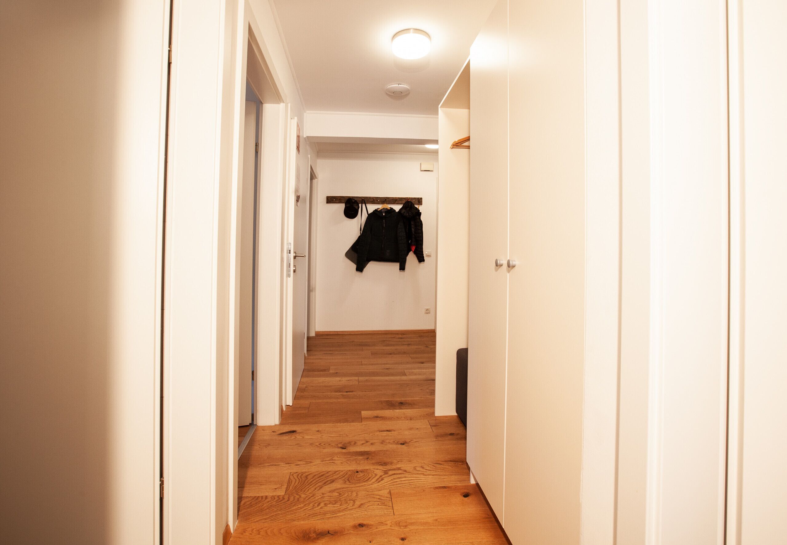 A narrow hallway with wooden flooring and white walls, leading to a room with a coat rack.