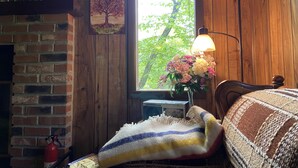 A cozy room with a brick fireplace, a wooden wall, a window with a view of trees, a bedside lamp, and a bedside table with a vase of flowers. - 4br 2ba Chalet in Poconos that sleeps 10 (Bushkill)