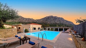 Un espace piscine aménagé avec des chaises longues et une table, entouré de murs en pierre et offrant une vue sur les montagnes en arrière-plan.