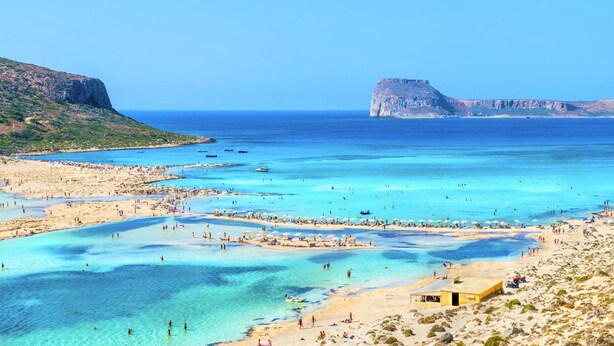 Une plage aux eaux turquoise et limpides, une falaise rocheuse sur la gauche et une grande formation rocheuse au loin.