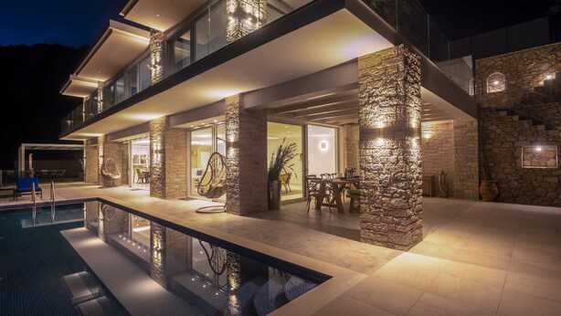 Uma casa moderna de vários andares com piscina, pilares de pedra e área de refeições com mobiliário externo.