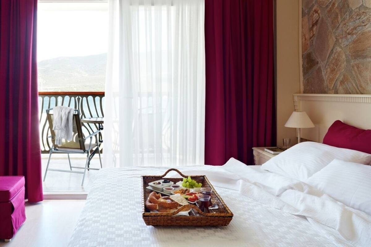 A hotel room with a bed, a tray of breakfast items, a balcony with a chair, and a painting on the wall.
