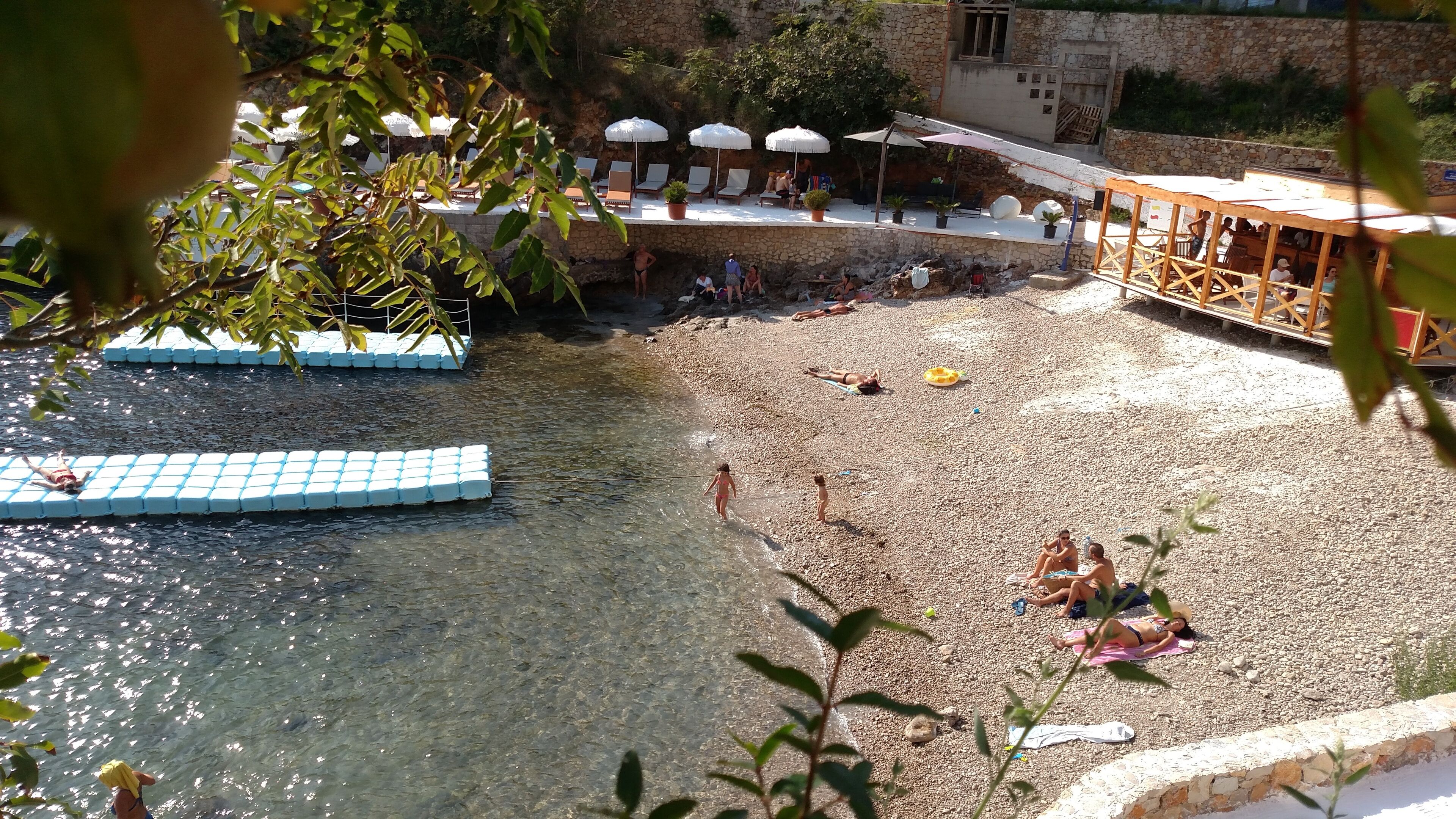 A pebble beach with clear water, lounge chairs, umbrellas, and a wooden structure.