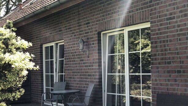 A brick building with large windows, outdoor seating with chairs and a table, and a small tree to the left.
