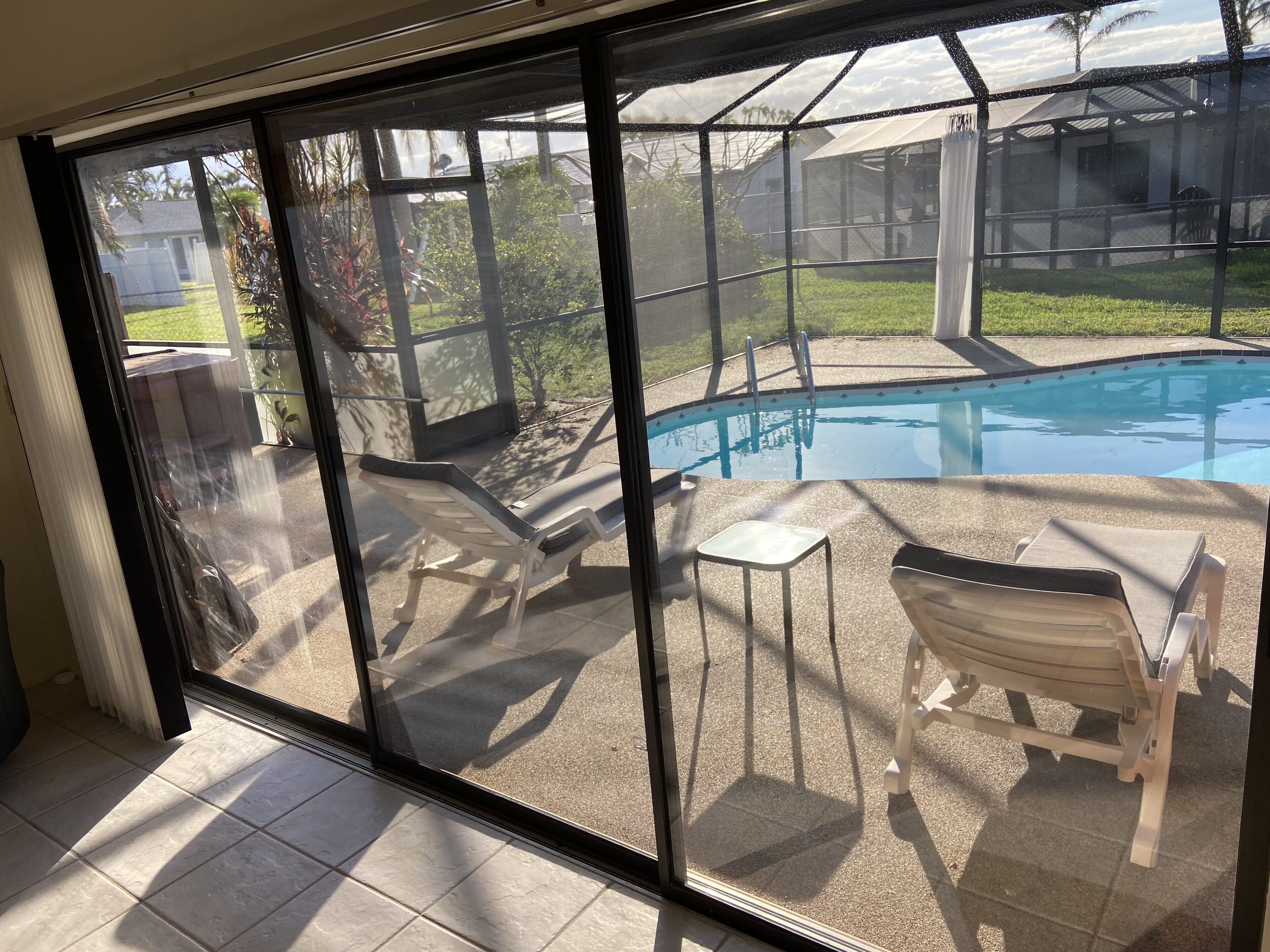 Zona de piscina con sillones de descanso y una pequeña mesa, vista a través de una puerta de cristal.