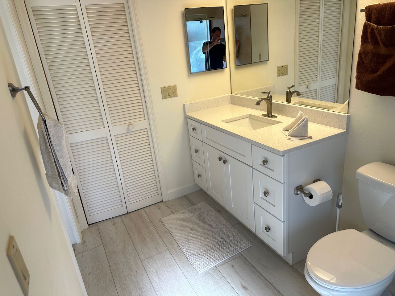 A bathroom with a white sink, mirror, and toilet. There is a towel hanging on the wall and a mat on the floor.