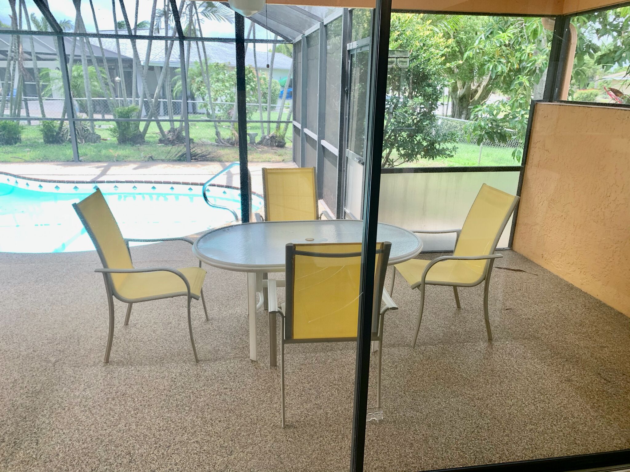 Un patio techado con una mesa redonda y cuatro sillas, con vista a una piscina y follaje.