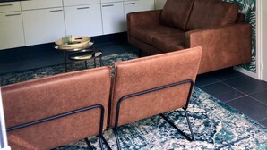 A compact kitchen with white cabinets, a black countertop, and a window. There is a brown sofa and two chairs in the living area. - Compact, trendy furnished, vacation bungalow. Enclosed garden. Pets welcome! (Bruinisse)