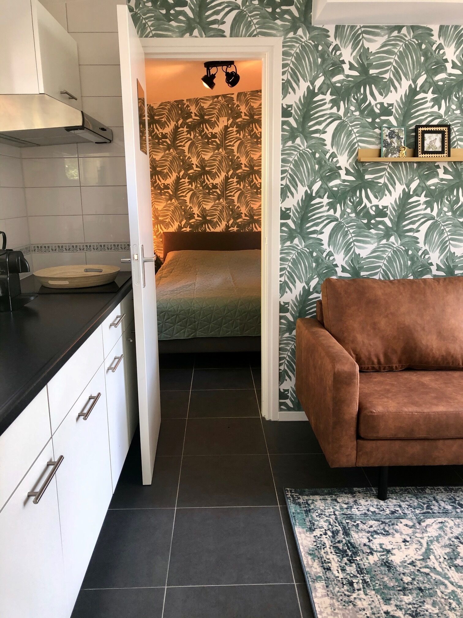 A compact living space with a kitchenette, a bed with a green quilt, a brown sofa, and a patterned rug.