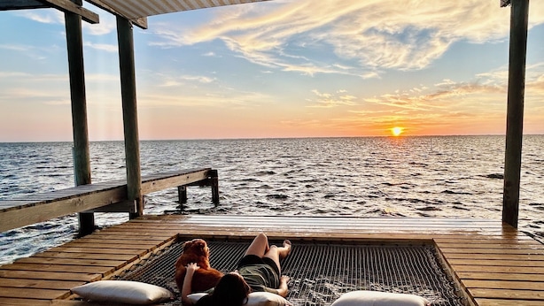 Eine Holzterrasse mit Blick auf den Sonnenuntergang über dem Meer, inklusive Grill aus Metall und Holzsäulen.