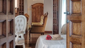 Una habitació amb una porta de fusta, una cadira tapissada de blanc, un butac armchair estampat i un llit amb una colcha blanca i un flòleg vermell.