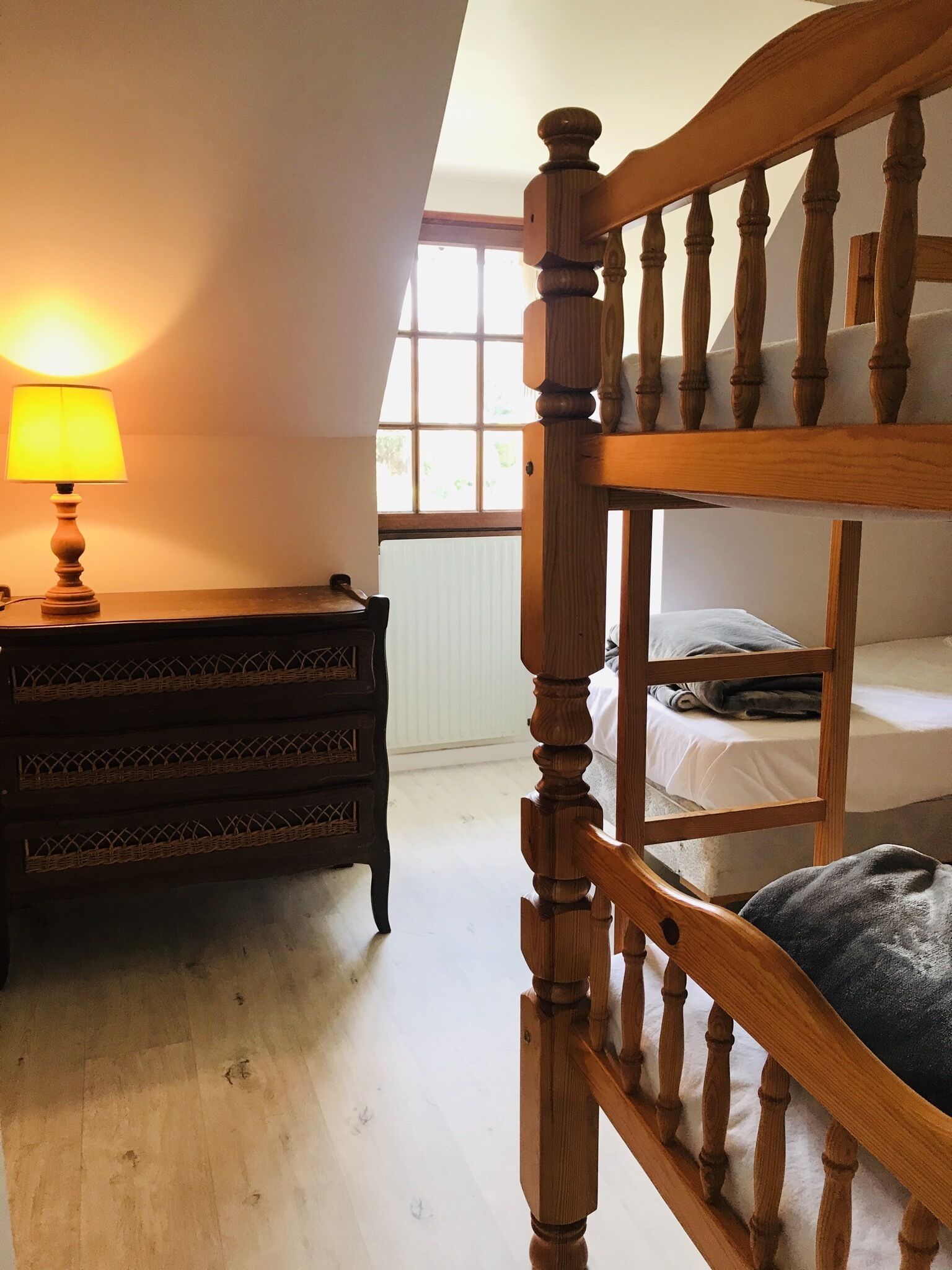Ein Zimmer mit einem hölzernen Etagenbett, einem Nachttisch mit Lampe und einem Fenster mit Blick auf Grünfläche.