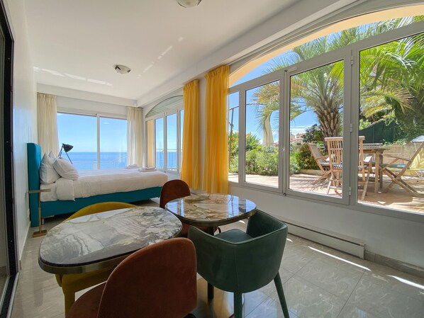 A room with a bed, a table, and chairs, large windows with curtains, and a view of the sea.