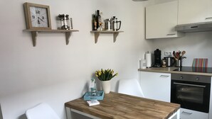 Eine kleine Küche mit Holztisch, weißen Stühlen, einer Uhr und einer Kaffeemaschine auf einem Regal.