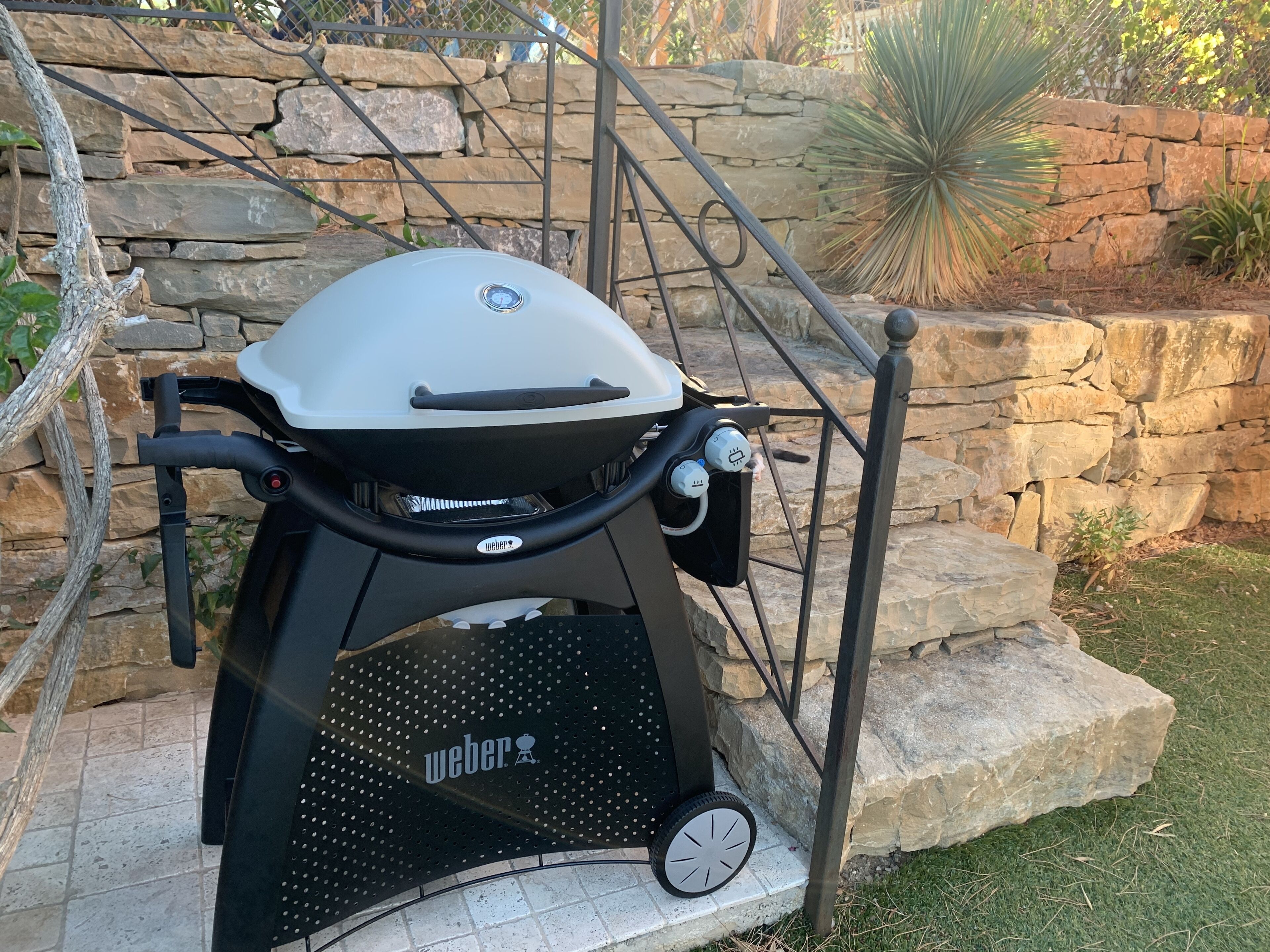 A portable Weber grill with wheels, set against a stone wall and surrounded by greenery.